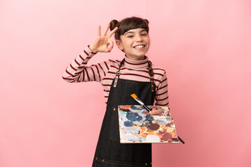 Little artist girl holding a palette isolated on pink background showing ok sign with fingers