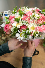 Close-up of the hands of a young female florist packing a beautiful composition of delicate pink roses, carnations and white daisies into a cardboard box and paper on the table. Vertical photo