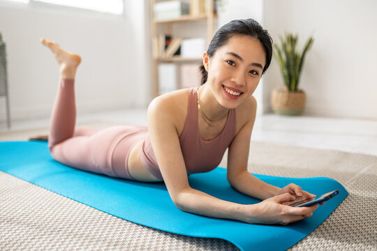 Smiling young asian woman holding mobile phone while relaxing after exercising at home. Female fitness girl looking at camera lying on mat. Sport, technology and healthy lifestyle concept.