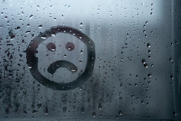Sad smiley drawn by hand on wet fogged glass from the rain, copy the space. Concept of loneliness,...