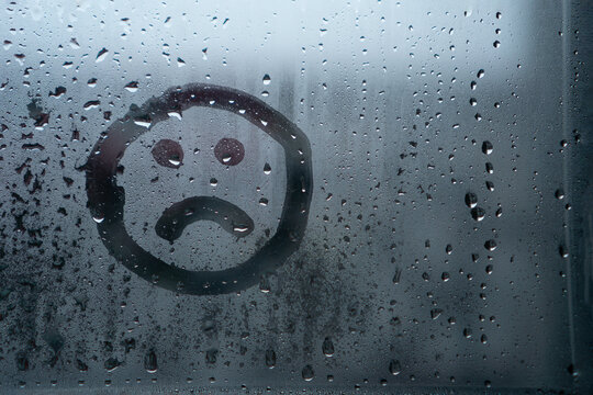 Sad smiley drawn by hand on wet fogged glass from the rain, copy the space. Concept of loneliness, sadness, bad weather