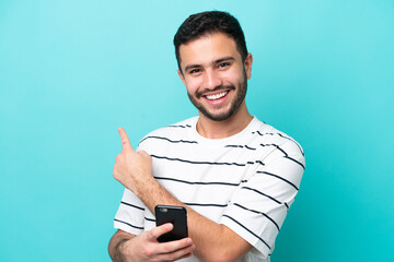 Young Brazilian man isolated on blue background using mobile phone and pointing back