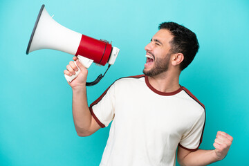 Young Brazilian man isolated on blue background shouting through a megaphone to announce something in lateral position