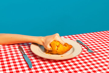 Female hand tasting crispy croissant on plaid tablecloth isolated on bright blue background. Vintage, retro style interior. Food pop art photography.