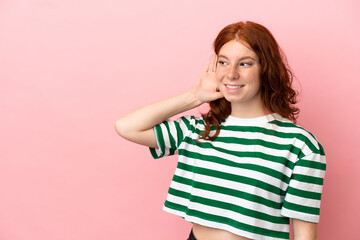 Teenager redhead girl over isolated pink background listening to something by putting hand on the ear