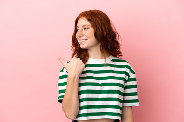 Teenager redhead girl over isolated pink background pointing to the side to present a product