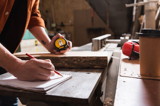 cropped view of furniture designer holding tape measure and writing in notebook.