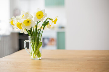  Kithen interior. Spring yellow flowers in a vase on the kitchen  table. Bouquet of daffodils in a transparent glass. The atmosphere of comfort and coziness. Selective focus. Copy space for text