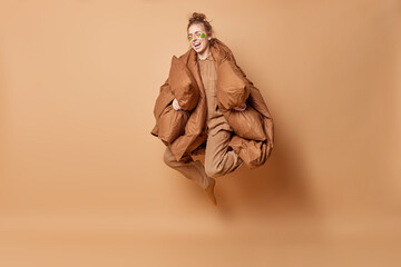 Overjoyed woman with hair bun smiles broadly jumps high feels glad dressed in nightwear covered with blanket holds two pillows isolated over beige background. People rest and sleeping concept