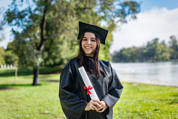Happy caucasian graduated girl with long brown hair. She is wearing a bachelor gown and a black mortarboard. She is holding a diploma - obrazy, fototapety, plakaty