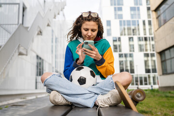 One young caucasian woman female sitting on the bench in front of the building or at stadium with soccer ball waiting for the football game using mobile phone real people copy space