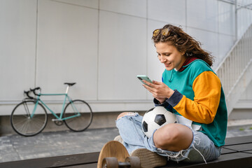 One young caucasian woman female sitting on the bench in front of the building or at stadium with soccer ball waiting for the football game using mobile phone real people copy space