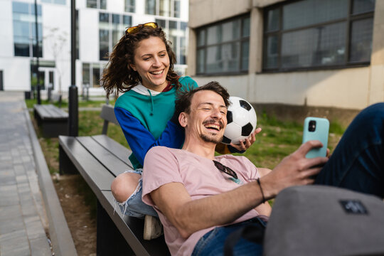 Couple woman and young caucasian man modern happy adult male and female boyfriend and girlfriend smile sitting in front of stadium holding soccer ball outdoor in day football game concept copy space - Powered by Adobe