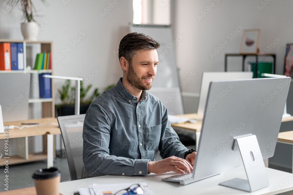 Poster Cheerful middle aged businessman sitting at worktable, typing on computer keyboard, working on marketing research