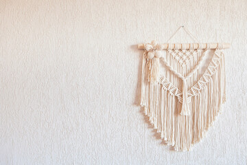 Handmade macrame wall decoration with wooden stick hanging on a white wall. Macrame braiding and cotton threads. Female hobby. ECO friendly modern knitting natural decoration in interior. Copy space