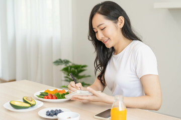 Obraz na płótnie Canvas Diet, Dieting asian young woman write diet plan right nutrition on table with fresh vegetables salad, almond is different food ingredients in green. Nutritionist of healthy, nutrition of weight loss.