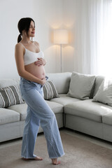 Young beautiful woman on second trimester of pregnancy. Pregnant female wearing supportive seamless maternity bra and unzipped mom jeans with arms on a belly. Expecting a child. Background, copy space