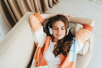 Young relaxed woman is laying down on sofa and  listening to a music with headphones on her ears.