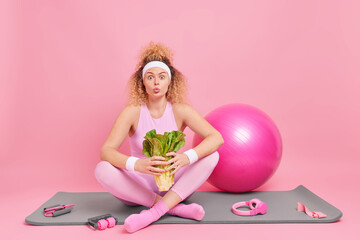 Obraz na płótnie Canvas Sporty woman with curly hair dressed in activewear holds green vegetable poses on fitness mat with sport equipment leads healthy lifestyle isolated over pink background. Sporty lifestyle concept