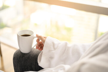 Side view of woman in bathrobe sitting near the window while holding white cup of coffee and enjoy wonderful morning in the hotel, detaching from work.