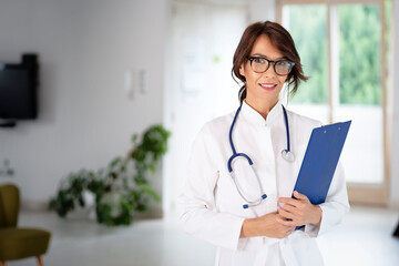 Smiling female doctor standing at hospital corridor while looking at camera and smiling