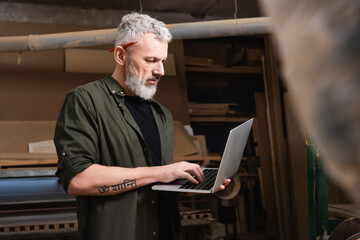 grey haired furniture designer in woodwork studio typing on laptop on blurred foreground.
