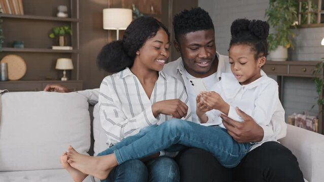 Happy african family parents with cute schoolgirl kid child relax on sofa using funny smartphone app reading joke online laughing having fun with phone together smile looking at mobile screen at home