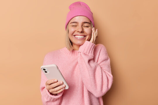 People and technology concept. Positive woman smils broadly keeps eyes closed waits for important call has glad expression touches face gently wears soft jumper isolated over brown background