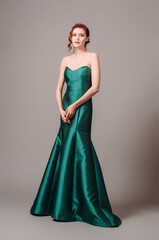 Fototapeta premium Emerald ball gown. Young lady in long green evening dress. Ginger woman posing in studio. Beautiful model wearing high heels, feminine look for an event. Women's classic fashion.