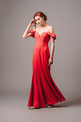 Elegant young lady in scarlet long dress with plunge neckline on high heels walking and smiling on grey studio background. Festive female gown.