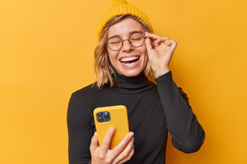 Joyful teenage girl watches funny video content via cellphone laughs happily keeps hand on rim of spectacles wears hat and black poloneck poses against yellow background checks positive email