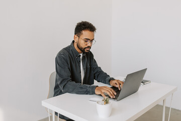 Side shot of smiling happy african male in stylish grey shirt with curly hair and eyeglasses working on laptop and typing while sitting at the white desk in airy cozy modern office studio 4k