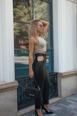 teen leisure. a young blonde woman in a beige top and black leather pants stands fashionably in profile and smiles sweetly near the  white street cafe background. sport lifestyle concept, free space