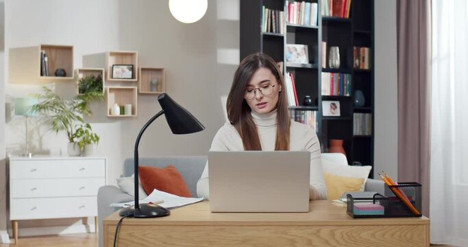 Caucasian young pretty woman in glasses sitting at desk, opening laptop computer and starting to work. Beautiful female freelancer start working day at home. Cozy room. Student studying online.