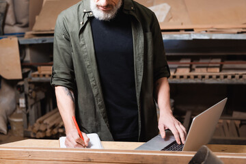 cropped view of furniture designer writing in notebook near laptop on workbench.
