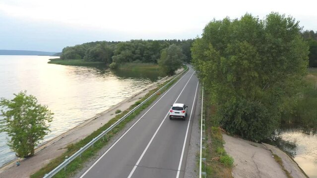 Aerial shot of car rides through road near lake. White SUV drives at dam route on summer day. Auto moves through bridge of river or pond. Scenic landscape way. Concept of travel and holidays. Top view