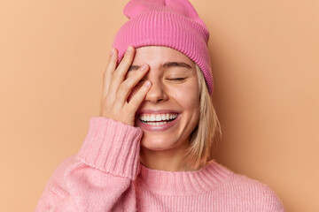 Happy woman makes face palm smiles broadly shows white teeth closes eyes from satisfaction feels very glad hears excellent news wears hat and sweater isolated over brown background. Emotions concept