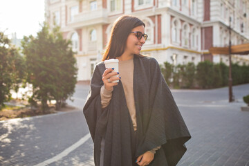 Elegant press woman in grey poncho and eyeglasses is drinking coffee and looking aside on sunny street 