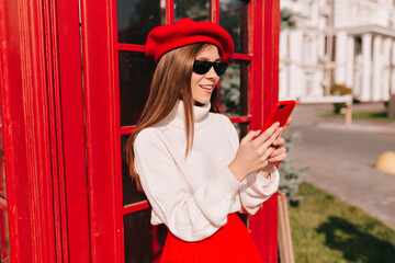 Happy lovely stylish girl with light-brown straight hair is wearing red beret and white pullover in sunglasses is using smartphone on sunny street