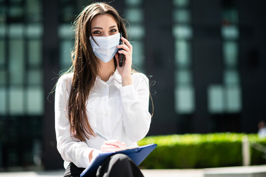 Smiling businesswoman writing a document outdoor while on the phone and wearing a protective mask against covid 19 coronavirus pandemic