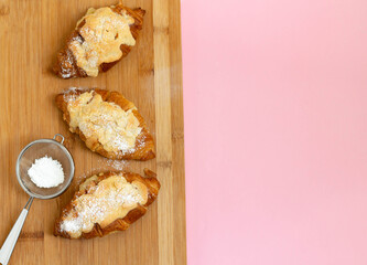 Croissants with almond cream and powdered sugar on a wooden board on a pink background. View from above