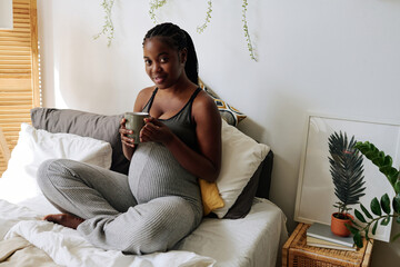 Portrait of African pregnant woman smiling at camera while enjoying tea sitting in bed
