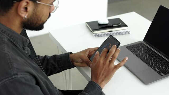 Black male person sitting at the white table in front of laptop holding cell phone scrolling and tapping screen of mobile phone, looking at stock market etf price in light office studio 4k