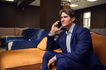Portrait of a handsome mature man in business suit, client customer buyer talking on mobile phone while visiting a furniture store and testing sofa. Sales manager, interior designer at work