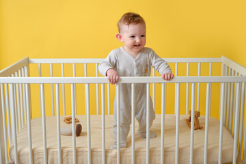 Happy infant baby boy is standing in a crib, yellow studio background. Smiling child in white...