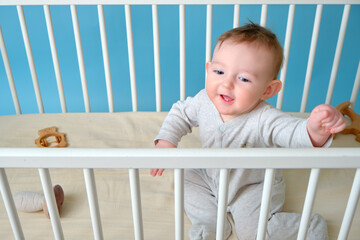 Happy infant baby boy is sitting in a crib, studio blue background. Smiling child in white pajamas, copy space