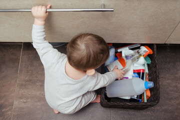 Toddler baby boy is playing in the toilet room with cleaning detergents. Child plays on a brown floor in a beige bathroom with household chemicals