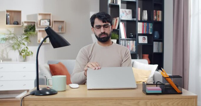 Arabian young male freelancer finish working day at laptop computer, taking off glasses and resting as having headache. Tired after workday in front of screen. Ending job. Arab busy male end work.
