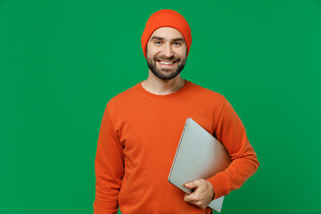 Young smiling programmer student cheerful happy man 20s in orange sweatshirt hat hold closed laptop pc computer look camera isolated on plain green background studio portrait People lifestyle concept