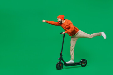 Full body side view young smiling happy fun man 20s wear orange sweatshirt hat riding electric scooter do super hero fly gesture isolated on plain green background studio. People lifestyle concept © ViDi Studio